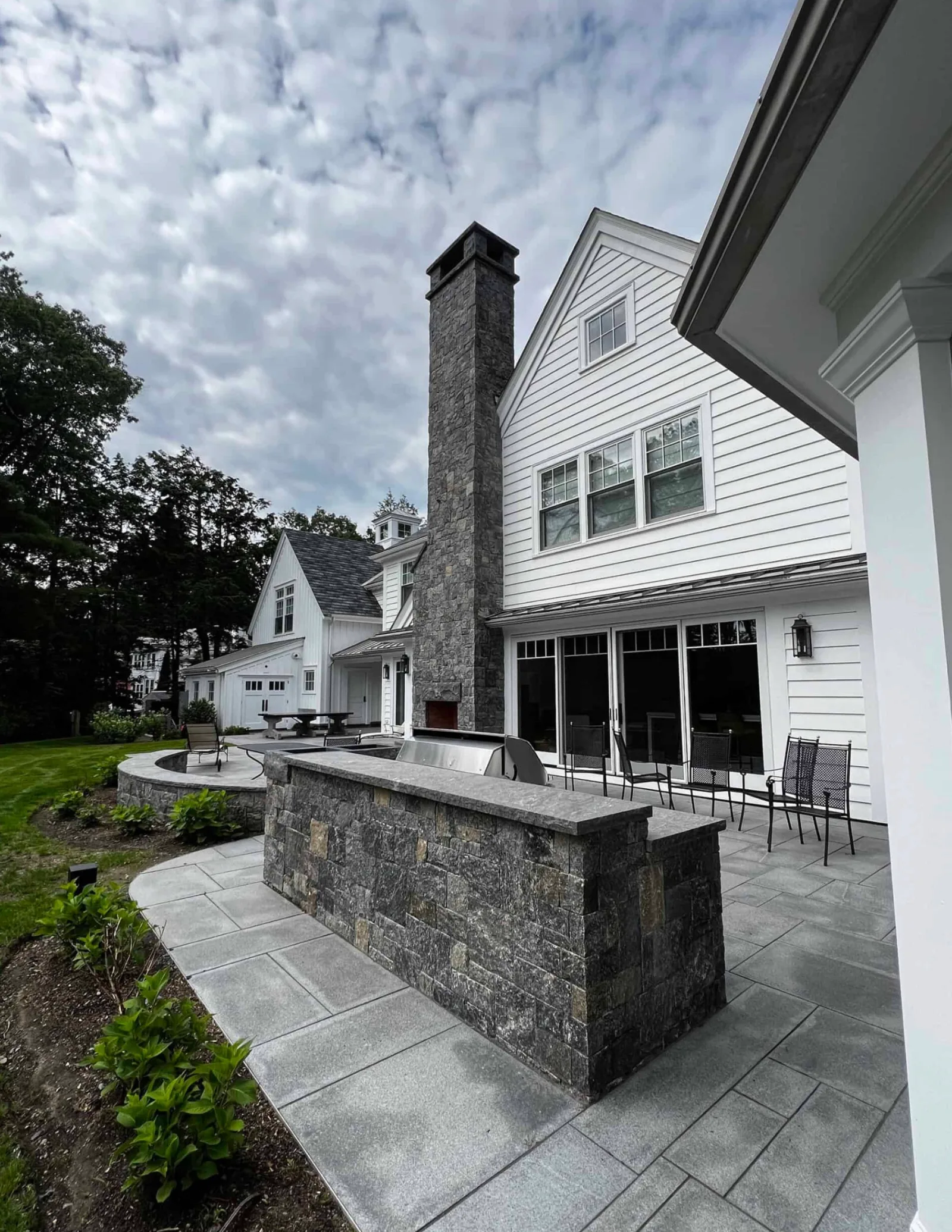 Stone-clad outdoor grill island on a paved patio beside a tall stone chimney