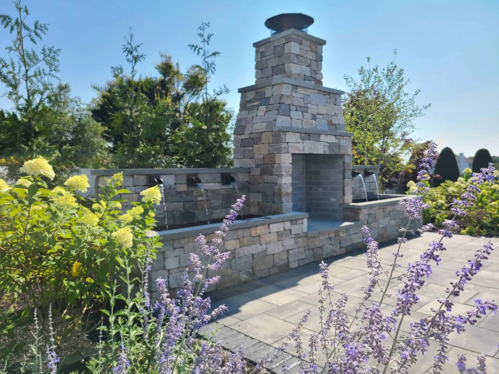Outdoor stone fireplace with water features on both sides beside a paved patio and flowering plants